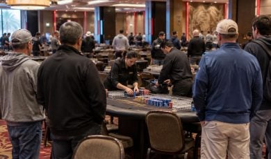 A casino dealer sorts poker chips at a table while players watch in a large, dimly lit gaming hall.