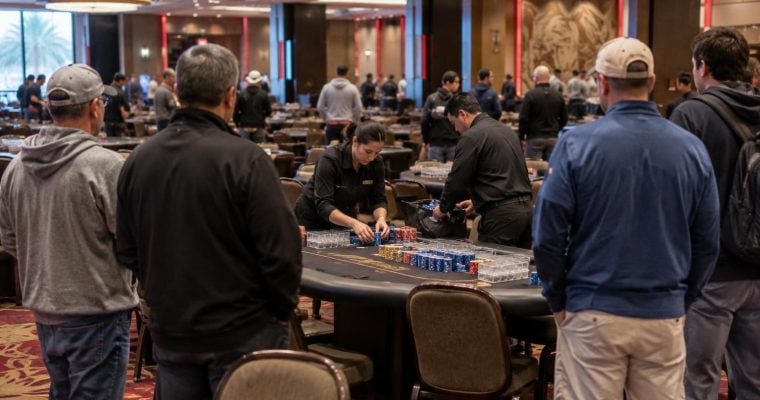 A casino dealer sorts poker chips at a table while players watch in a large, dimly lit gaming hall.